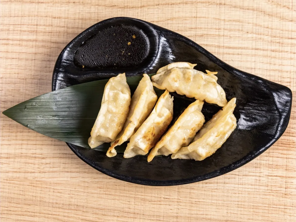 Six pan-fried chicken gyoza arranged on a bamboo leaf beside a dish of gyoza sauce