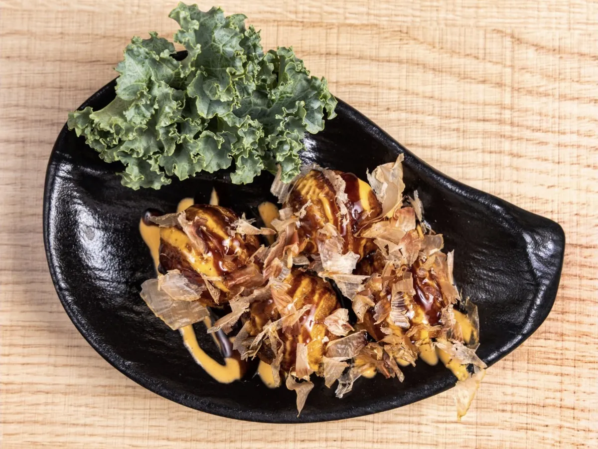 Takoyaki drizzled with sauce and mayonnaise under a heap of bonito flakes, served in a black dish with kale